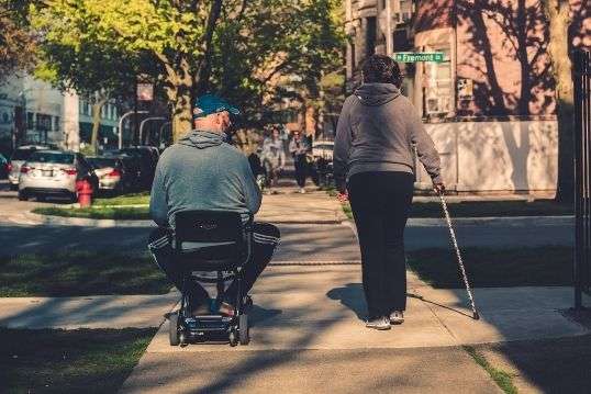 un homme et une femme traverse la rue avec un fauteuil roulant et une canne